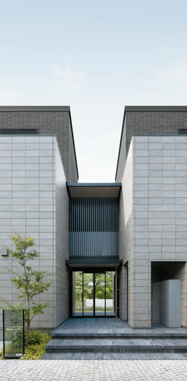 端正な箱型の建物にアクセントを添えるエントランス。ダーク系の仕上げが奥行きを生む。
